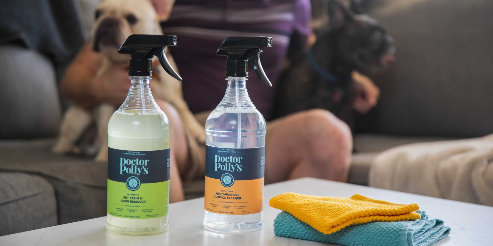 A bottle of Dr. Polly's Pet Stain & Odor Remover and a bottle of Dr. Polly's All Purpose Oxy Cleaner on a table next to cleaning rags. Two French Bulldogs in the background sitting on a couch.